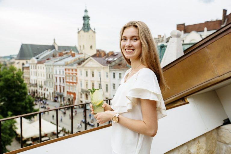 Happy blond smiling woman holding a glass of some drink while enjoying the european city view. Relaxing, tourism and resting concept.