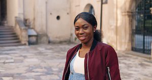 Portrait of charming African American young stylish woman looking and smiling to camera while standing in antique ruins in town. Outside. Beautiful joyful female tourist at nice architecture.